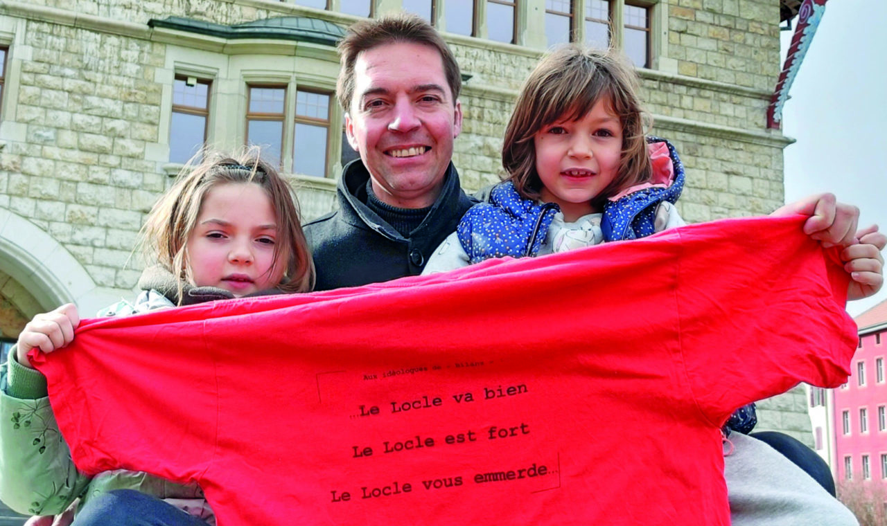 « Le Locle  vous emm… » Cédric Dupraz présente avec ses filles Camille et Elsa (à droite), le tee-shirt sorti à l’époque  « pour les idéologues de Bilanz ». (dr)